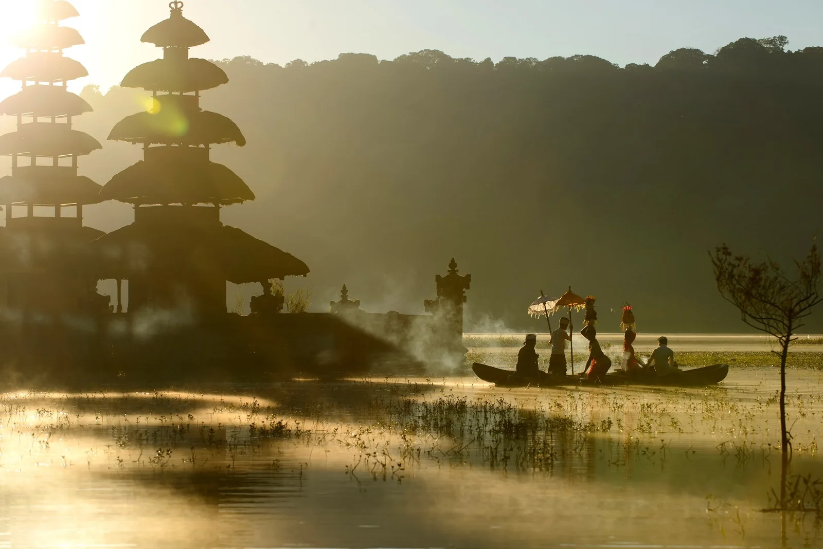 A boat carrying people with a ceremonial umbrella crosses a misty lake beside tiered temple shrines at sunrise in Bali.