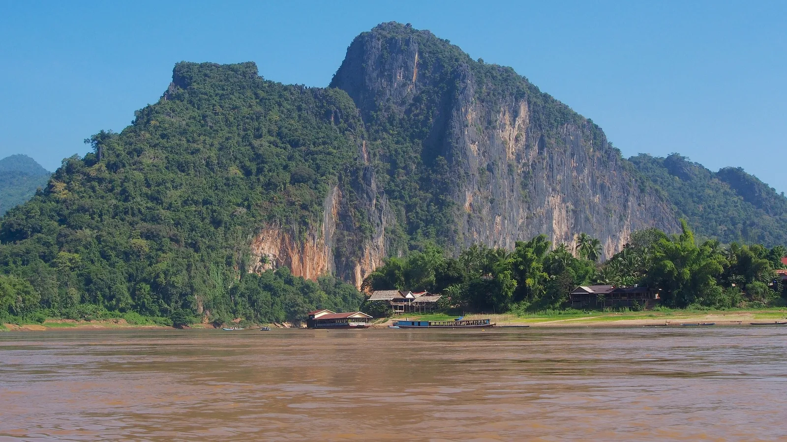 A limestone karst mountain rises above a riverside village along the Mekong River near Luang Prabang.
