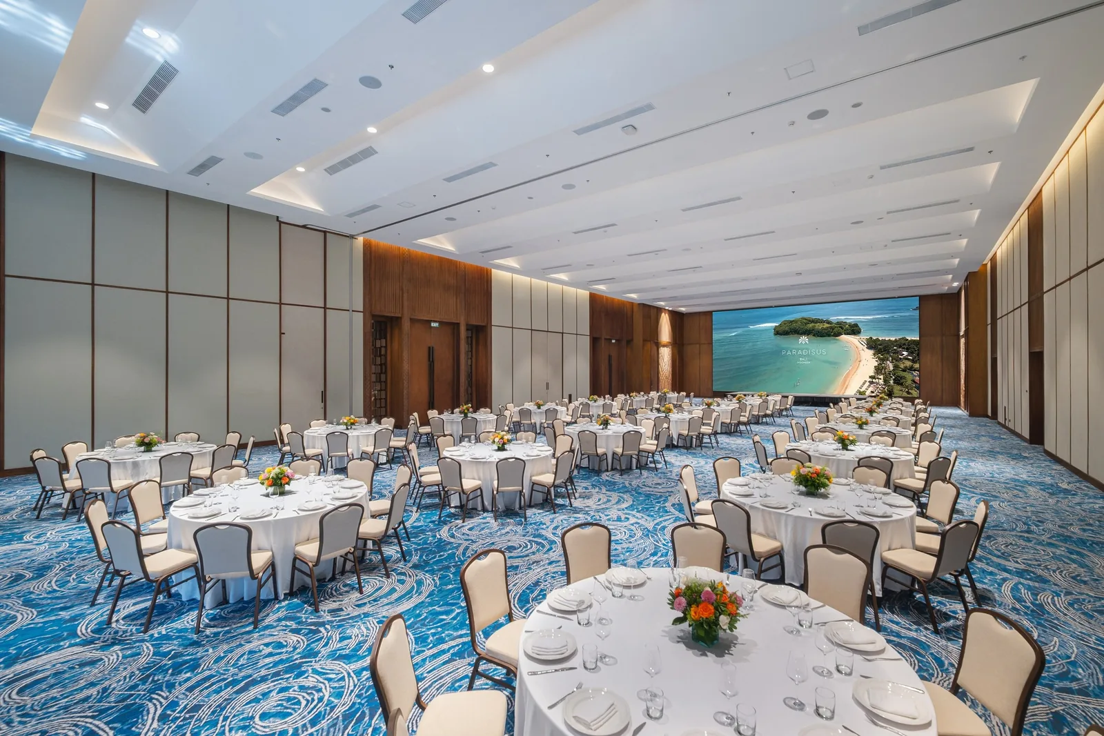 A grand ballroom at a Bali hotel set with round tables for a banquet and a large screen showing a beach view.