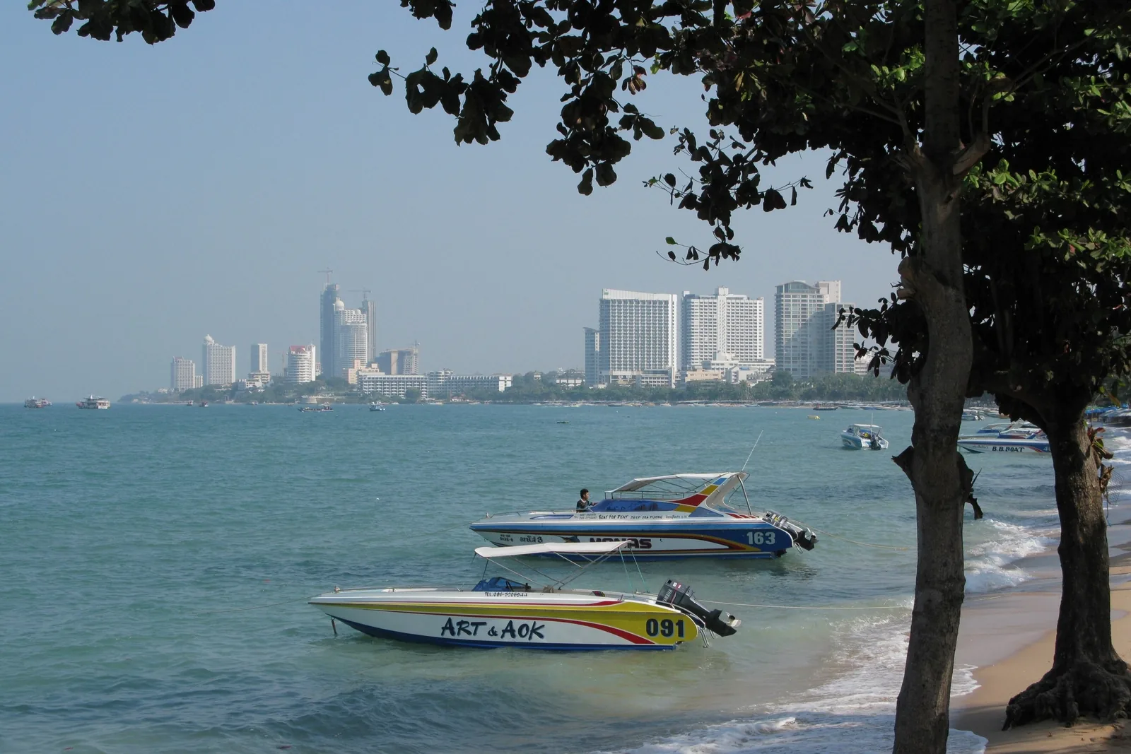 Speedboats float near the shore of Pattaya Beach with the city skyline visible across the bay.