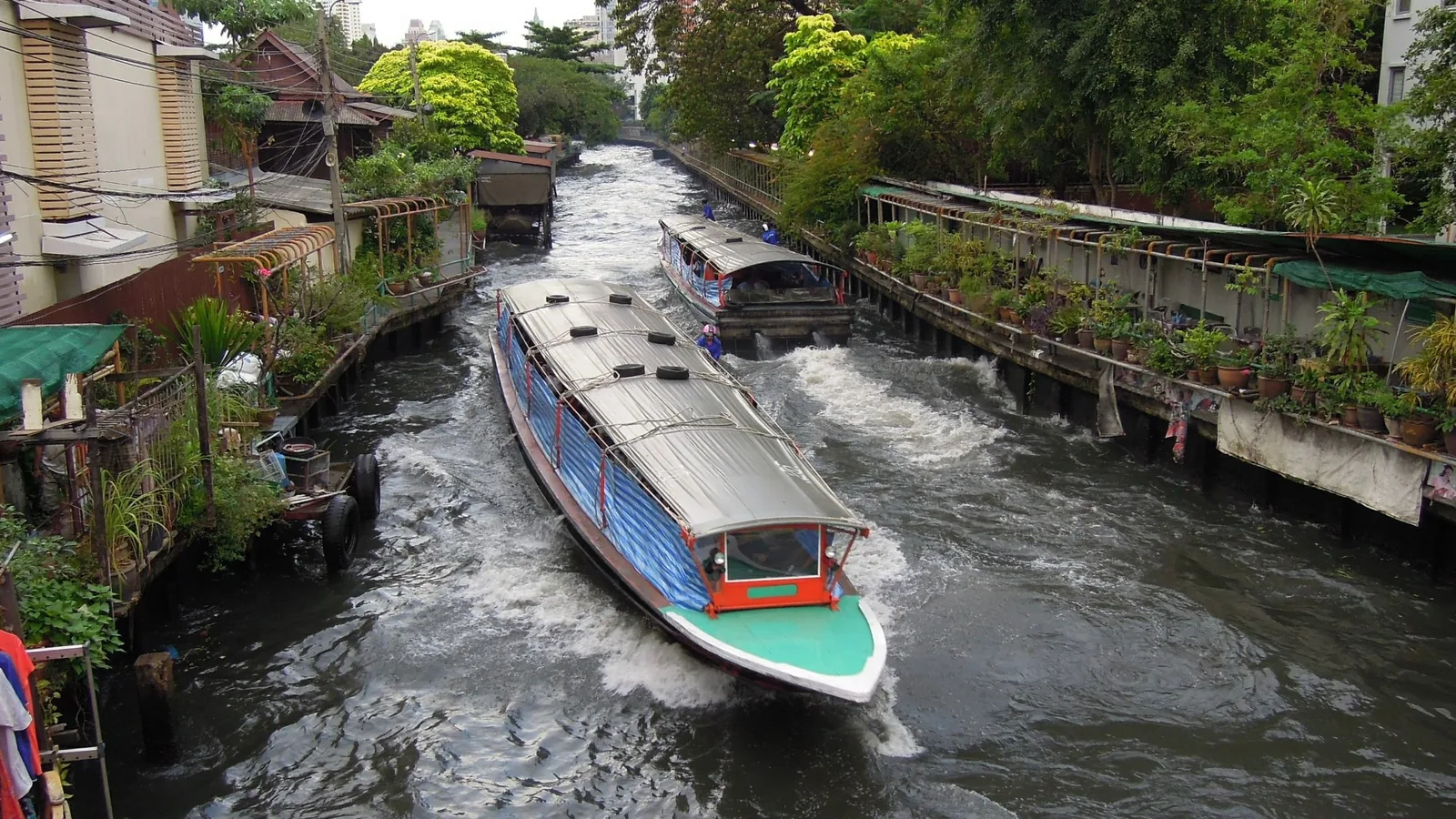 Long-tail express boats speed along Khlong Saen Saep canal lined with buildings and greenery in Bangkok.