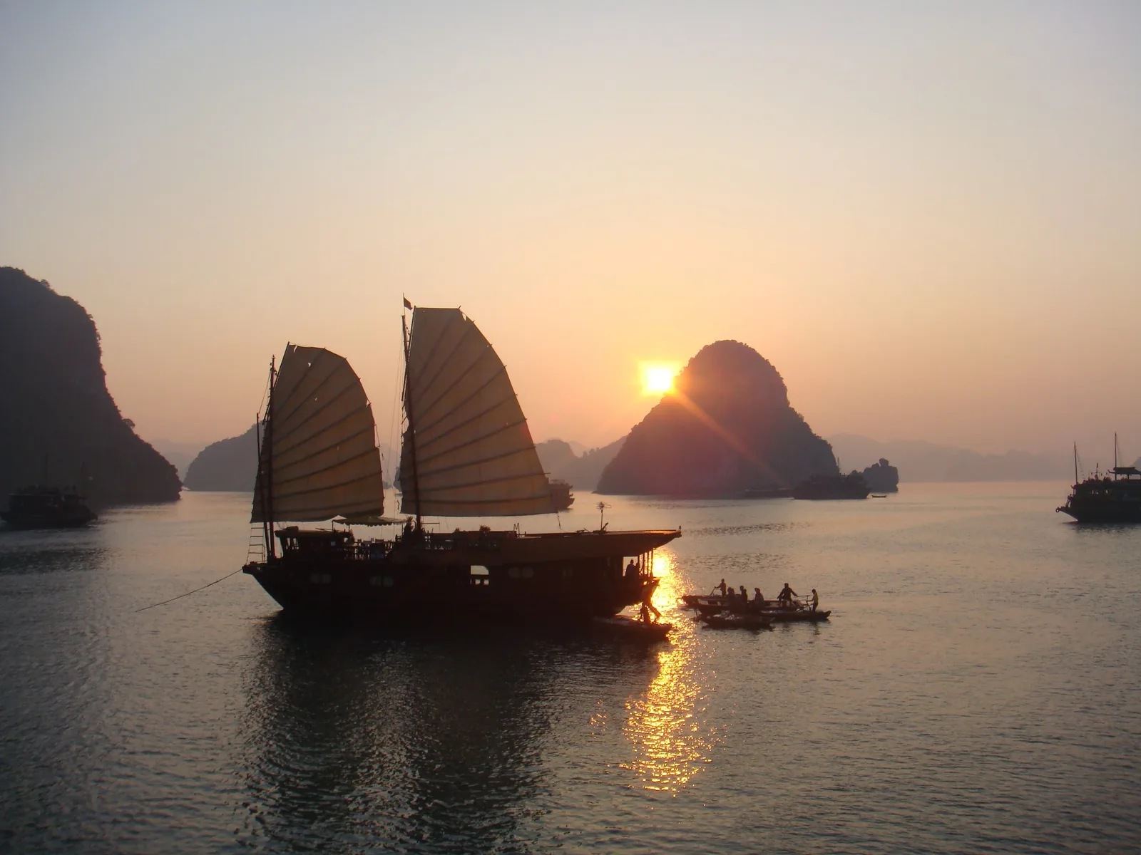 A traditional junk boat with sails silhouetted against the sunset among limestone karsts in Ha Long Bay.