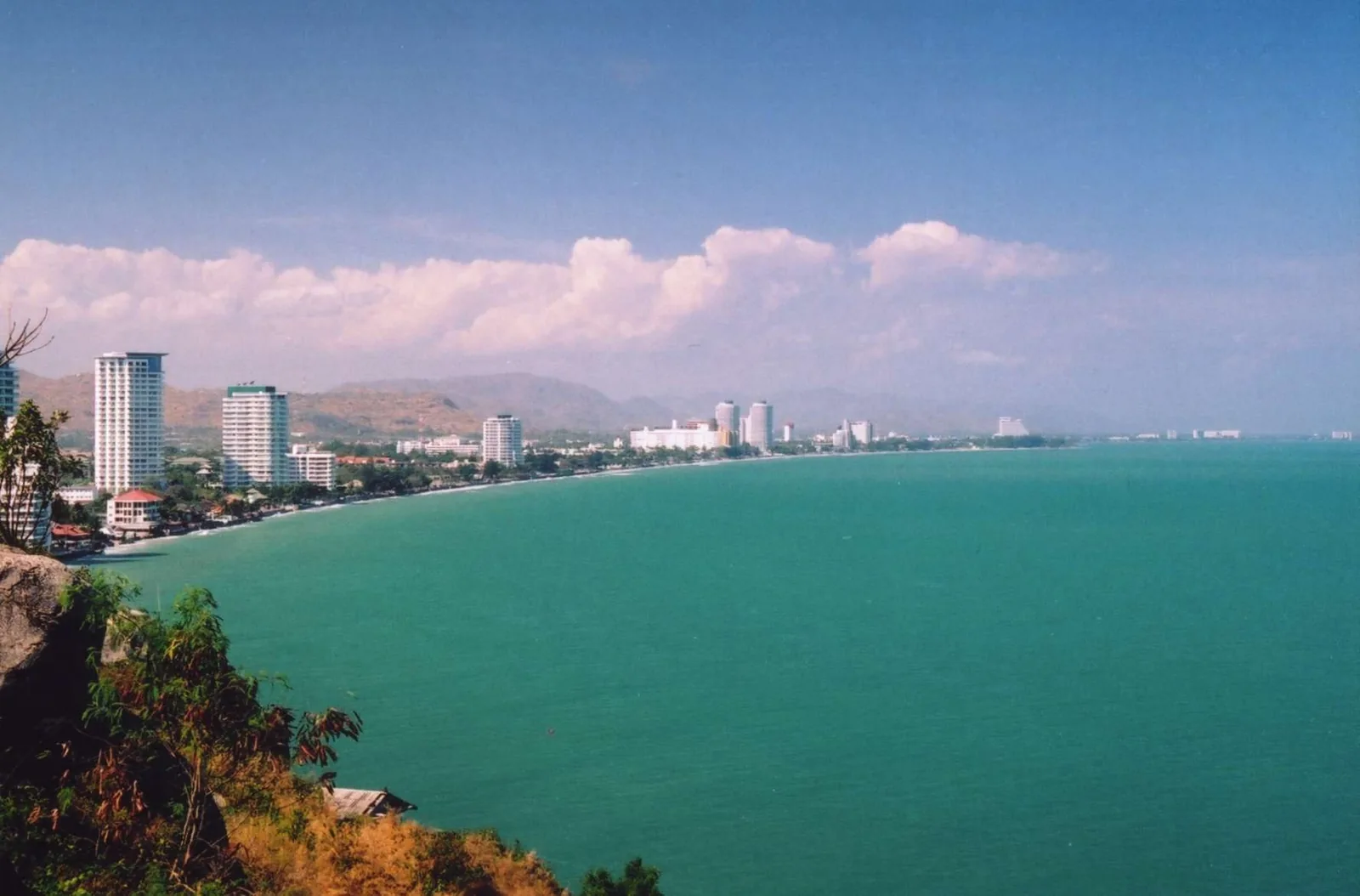 A curved coastline of Hua Hin with beachfront hotels and mountains beyond turquoise sea, seen from a hilltop viewpoint.